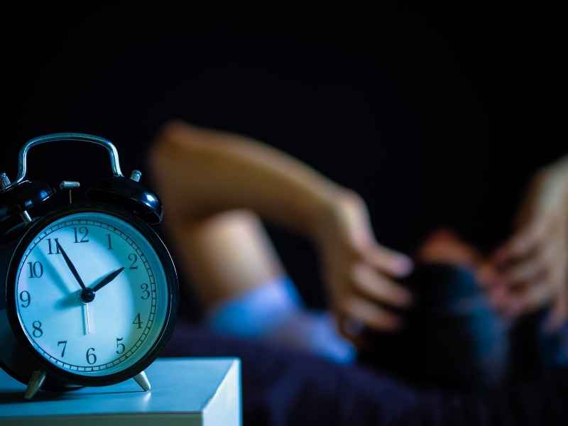 reloj sobre una mesita y al fondo una persona que sufre de insomnio frecuente